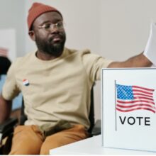 A black man who uses a wheelchair puts his ballot in a box that says vote and has an American flag on it