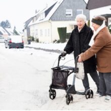 A woman assists an older woman using a walker in crossing a snow street