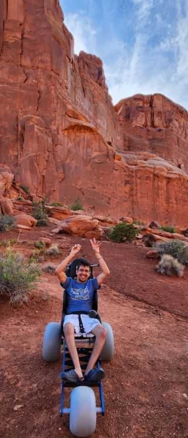 A young man looking excited sitting in his all-terrain wheelchair in front of a beautiful red rock formation