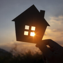 A hand holds a cutout of a house in front a sunset so that the sun's rays come through the houses windows.