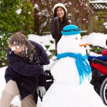 A group of three pre-teens take part in inclusive winter activities. The kids throw snowballs at each other in front of a snowman wearing a blue hat and scarf. The kids wear winter clothing and one child uses a wheelchair.